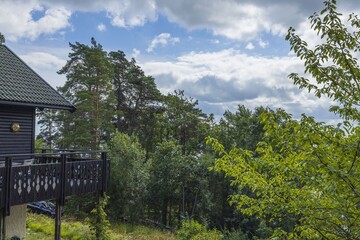 Naklejka premium Beautiful view of part facade of typical wooden Swedish house with wooden patio. Tops of green forest trees on blue sky and white clouds background on sunny summer day. 