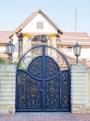 Rounded iron gates with a gilded wrought pattern and a fence enclosing a cottage