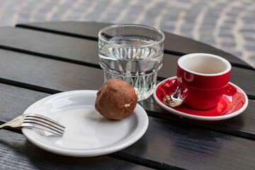 A small red cup with black coffee stands on a red saucer. There is a transparent glass of water nearby. A dark wooden table is located near a small cafe on a narrow street.