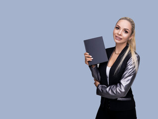 Portrait of a happy blonde girl in a dark jacket with a notepad in her hand, standing on a gray-blue background, looking at the camera and smiling.