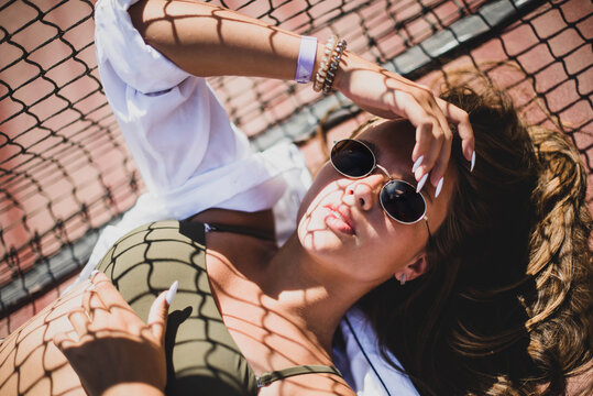Stylish cute teenage model dressed in summer hipster vintage white stylish clothes. Trendy girl posing in sunglasses lying on a tennis court. The original shadow from the grid at the sports field