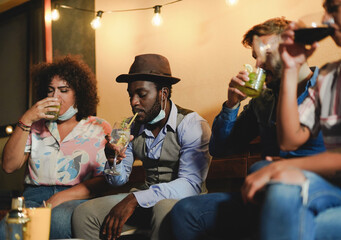 Multiracial friends drinking cocktails at home outdoor on patio while wearing protective face mask under chin