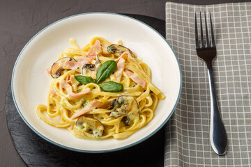 a plate of pasta with ham and mushrooms in a creamy sauce with basil on a black round stand with a fork on a napkin.