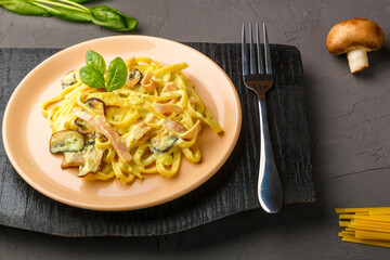 pasta with ham and mushrooms in a creamy sauce on a black board with a fork next to champignons and pasta.