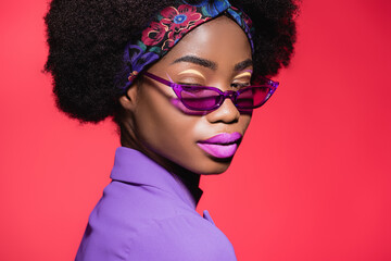 african american woman in purple stylish outfit and sunglasses isolated on red