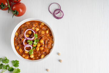Top view of chole masala or chana local indian street food made of chickpeas, tomatoes and cumin decorated with red onion rings and parsley served on white wooden background. Image with copy space