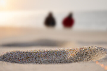 In the foreground is a macro shot of sand. In the background two people out of focus are walking along the shore