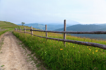 Fototapeta na wymiar Mountain landscape, road with a wooden fence along a bright meadow, silhouettes of mountains on the horizon, grey sky. Ukraine, Carpathians.