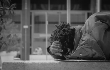 Grayscale shot of an African-American person wearing a coat and lying on a bench outdoors