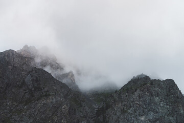 Dramatic bleak fog among giant rocky mountains. Ghostly atmospheric view to big cliff in cloudy sky. Low clouds and beautiful rockies. Minimalist scenery in mysterious place at early foggy morning.