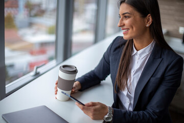 A young smiling businesswoman is looking through the window in the office with coffee and mobile in...
