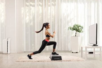 Young female exercising step aerobic in front of tv at home - obrazy, fototapety, plakaty