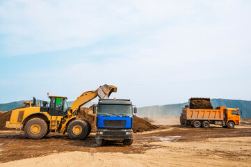 Obraz premium • Earthworks on a summer day in mountainous areas. Wheel loaders load soil into the bodies of dump trucks. Dust from dump trucks that transport this soil