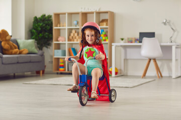 Cute smiling little girl in helmet and superhero red cape riding her tricycle at home