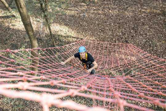 Well-equipped man climbing on a net outdoors, having active recreation in amusement park - Powered by Adobe