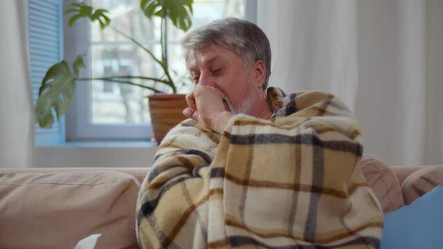 Elder sick man resting on sofa at home sneezing in paper tissue
