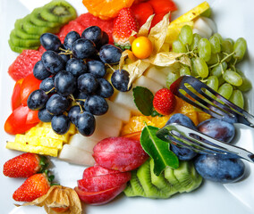 Assorted sliced fruit, black and white grapes, kiwi, prunes, pineapple, strawberries and physalis are on a platter. The view from the top.