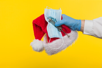 Close-up profile side view of a human hand in blue surgical gloves, holding a Santa Claus hat and a protective mask against COVID-19 coronavirus infections and an antibiotic syringe, 