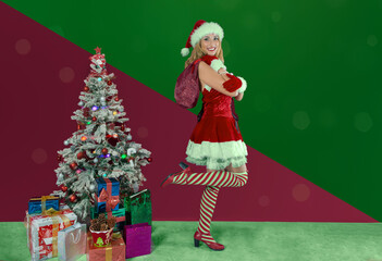 pretty young woman in santa claus dress next to the christmas tree full of gifts with wide smile