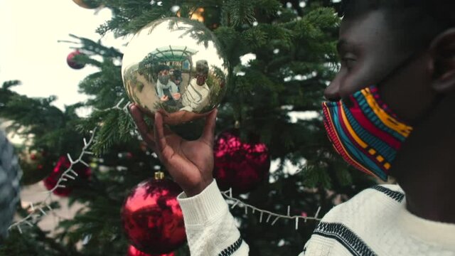 A masked African man is touching a New Year's toy at the Christmas tree. A playful mood with a girlfriend during Advent, a pre-holiday mood. Reflection in the ball.