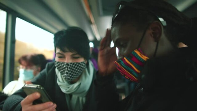 A black man shows pictures on the phone to a white woman on the bus in motion, scrolls with his finger on the screen. Communication and discussion of life stories. The faces are wearing masks.