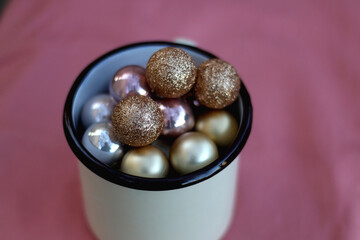 Enamel mug filled with gold baubles. Selective focus, pink background.