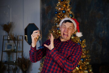 Young man in santa hat drinks champagne and grimaces from alcohol on background of Christmas tree in dark room. Concept of Christmas celebration at home.