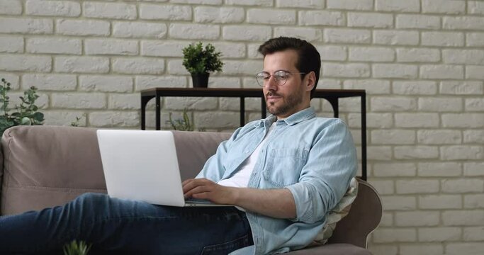 Thoughtful calm relaxed man in eyeglasses lying on comfortable couch, spending time online web surfing information, doing project research, working remotely from home, freelance workday lifestyle.
