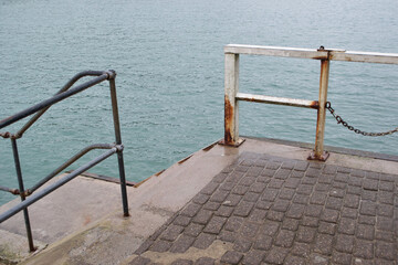 Rusty metal railings by the sea: white painted bars by a stone flagged pier on the coast, with steps going down to the water's edge