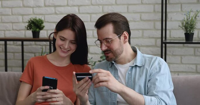 Head shot young man in glasses dictating credit banking card payment information to focused millennial woman, entering cvv code in mobile shopping app, happy couple involved in purchasing goods online