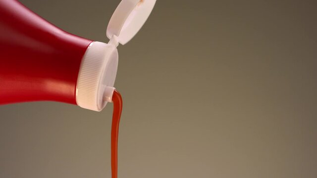 Close up of ketchup pouring from a bottle isolated on a beige background. Stock footage. Concept of cooking food, close up of red bottle of ketchup with white lid.