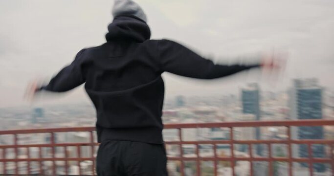 Male athlete warming up on roof. Sportsman in hat jumping and swinging arms during fitness workout on roof on cloudy day in city