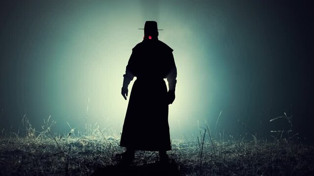 Silhouette of scary plague doctor man in long mantle costume standing in front of bright spotlight. Halloween, horror concept, historical medieval character