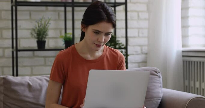 Happy millennial girl sitting on sofa with computer on aps, enjoying web surfing information, watching funny video photo content in social networks, communicating with friends or shopping online.