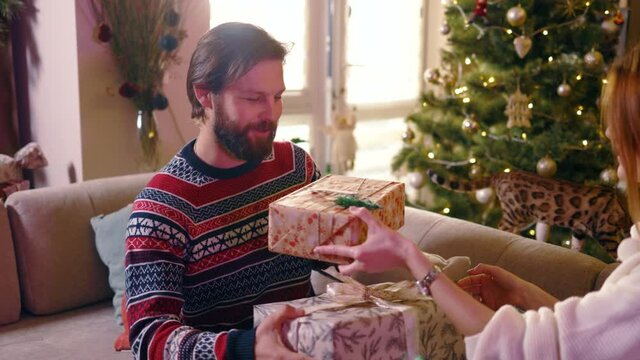 Happy caucasian couple exchanging gifts near christmas tree. Positive affectionate young family man and woman spending winter holidays at home.