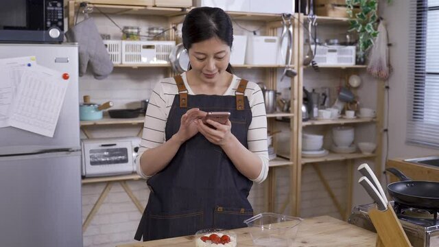 waist up female influencer wearing apron is food blogging by photographing her homemade cake with the smartphone in the kitchen.