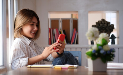 Young cheerful woman relaxing at her workspace and using smartphone browsing and messaging with friends.