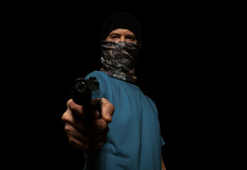 A criminal man in a mask on his face, aiming a gun at a camera, on a black background