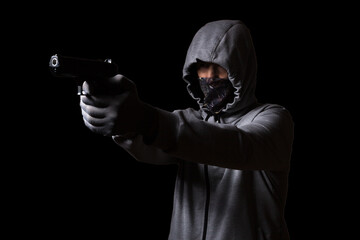 Criminal in a mask on his face and in a hood, aiming with a pistol, on a black background