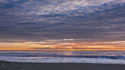 Obraz premium Sunset over the Black Sea. There are dense blue-white clouds in the sky. An orange glow over the horizon. There is a sunny path on the surface of the water. Foam from the waves on a pebble beach. 