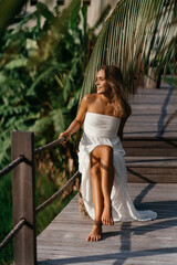 Fashion outdoor portrait of blonde woman, wearing elegant white dress, tropical leaves on background.