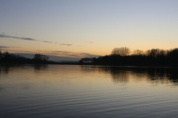 View at sunset with blue sky with some clouds at the horizon reflected on a lake in a cold autumn day, intense orange and dark colors at dusk in Stevenage, Hertfordshire, United Kingdom