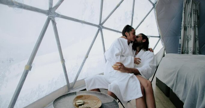 Beautiful woman sitting on a man's knee drinking white wine, in a white bathrobe and kissing. Attractive man and woman chilling in an igloo hotel room 4K