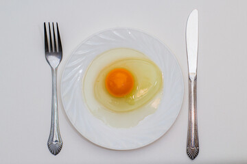 On a white plate, one raw egg, next to a fork and knife. Table setting. White background.