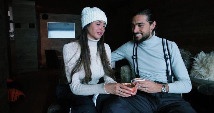 Attractive man and woman look at each other and kiss. The couple drinks coffee and hot chocolate after skiing, in front of a fireplace 4K