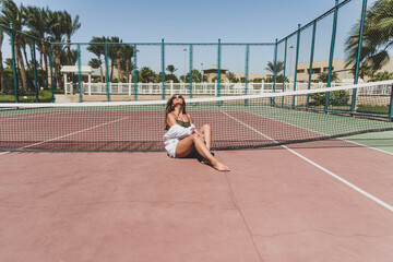 Stylish cute teenage model dressed in summer hipster vintage white stylish clothes. Trendy girl posing in sunglasses sitting on a tennis court. The original shadow from the grid at the sports field