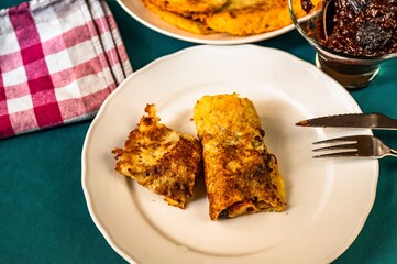 Wrapped fried potato pancake on white plate fork, plum jam and towel on green table.
