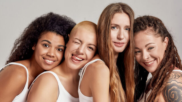 Close up portrait of four cheerful young diverse women, female friends smiling at camera while posing together isolated over grey background