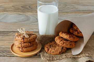 A stack of chocolate chips cookies on baking paper, with a glass of milk on a light wooden background
