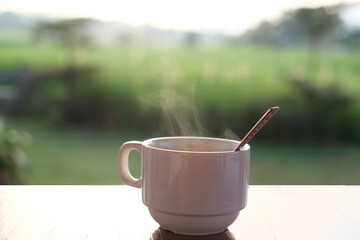 A white cup of hot coffee with steam on wooden table with sunlight morning. Green natural mountain and landscape background.  Lifestyle concept.
